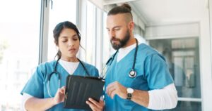 A male and female nurse in blue scrubs look at an iPad together