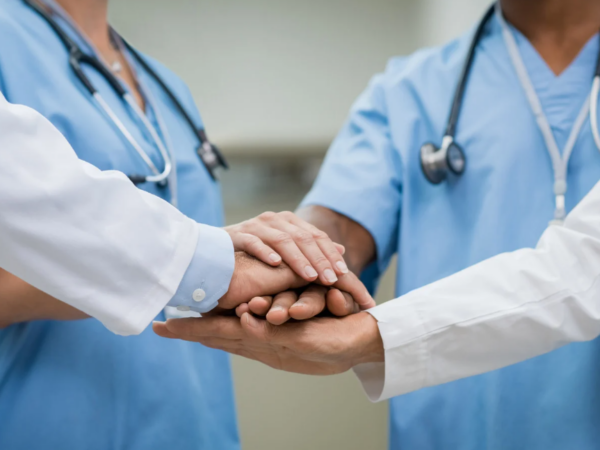A diverse group of nurses and interprofessional clinicians gathered around a table in a unit setting, collaborating, reviewing data, or in a shared governance-style meeting.