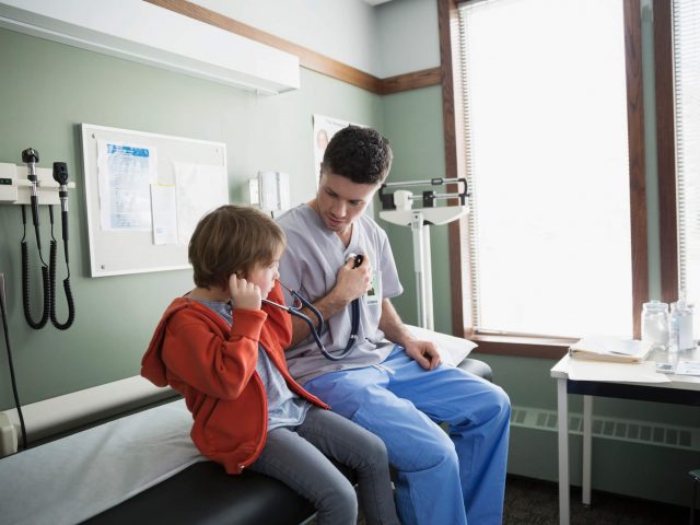 Male nurse sits on a patient bed with a little boy, letting the boy listen in his stethoscope