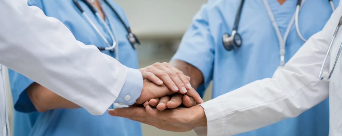 A diverse group of nurses and interprofessional clinicians gathered around a table in a unit setting, collaborating, reviewing data, or in a shared governance-style meeting.